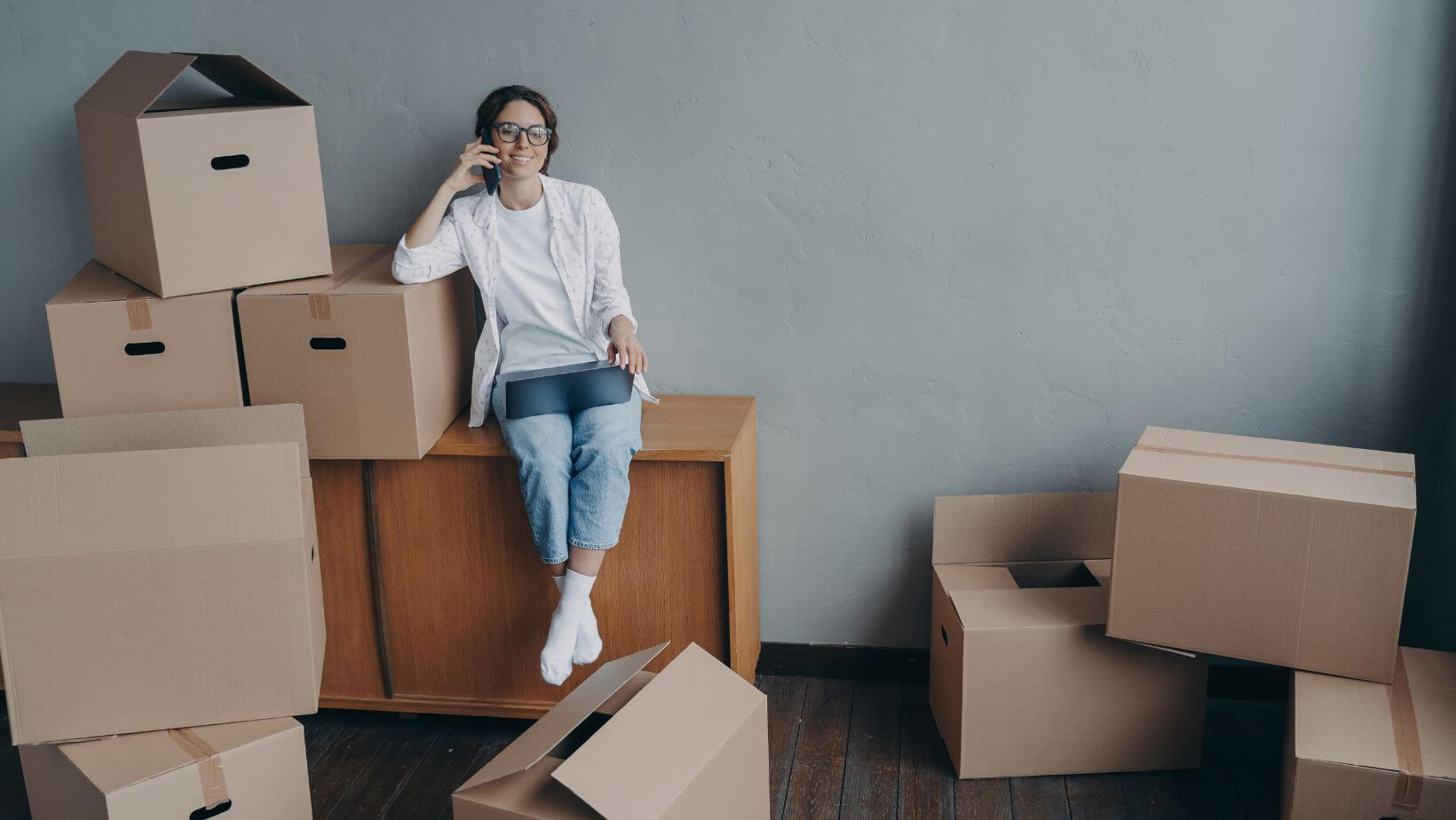 A woman on the phone, sitting on a dresser, surrounded by boxes.