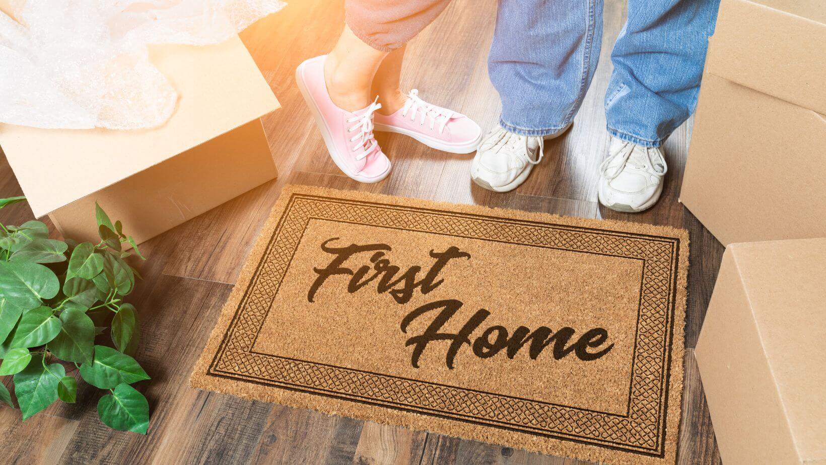 A couple standing in front of a doormat that says "First Home"