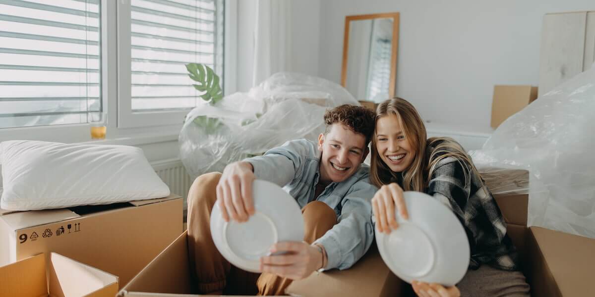 Young couple smiling while packing plates into boxes, showing how to pack dishes for moving without damage in a home relocation setting.