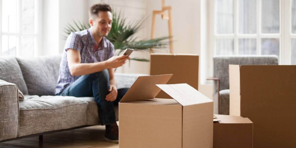 Man on couch with moving boxes checking phone, illustrating what is full service moving planning and coordination.