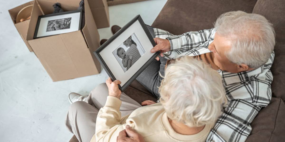 Elderly couple sitting on couch looking at framed photo while packing boxes, illustrating moving tips for seniors and downsizing process.