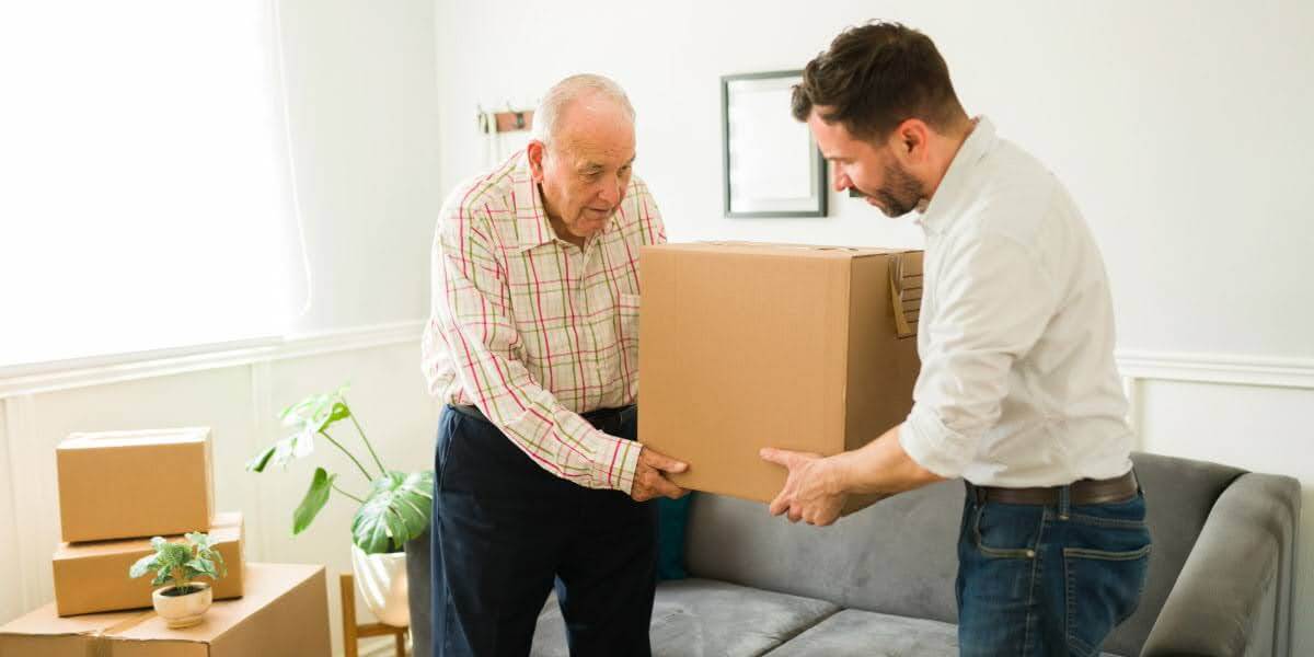 Younger man helping elderly man with a moving box in a home setting, illustrating moving to assisted living with family support.